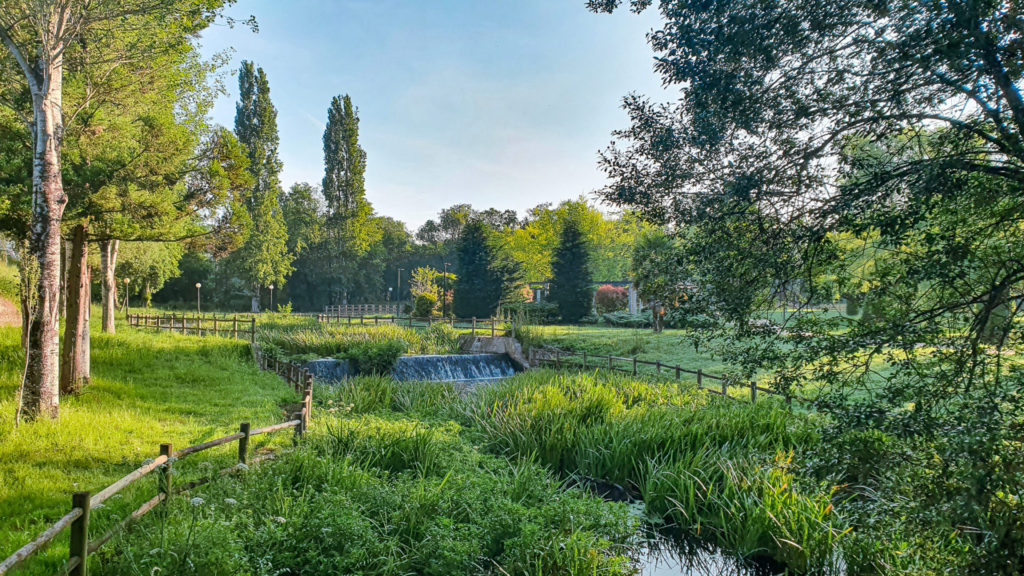 A park with a small river and a tiny waterfall among lush green trees on Via de la Plata