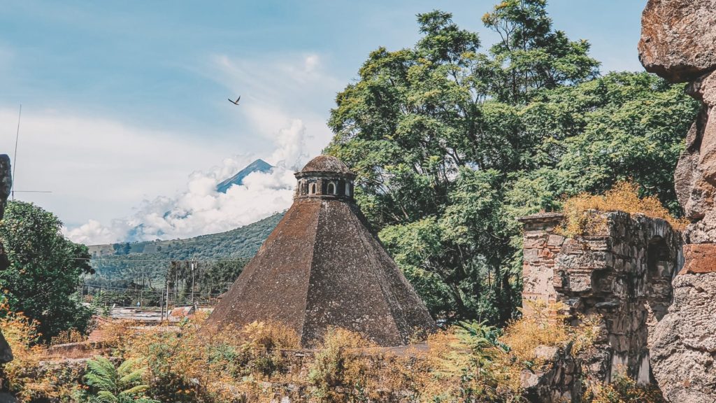 The roof of a colonial building, ruins in the bottom right corner and Fuego volcano surrounded by clouds in the background