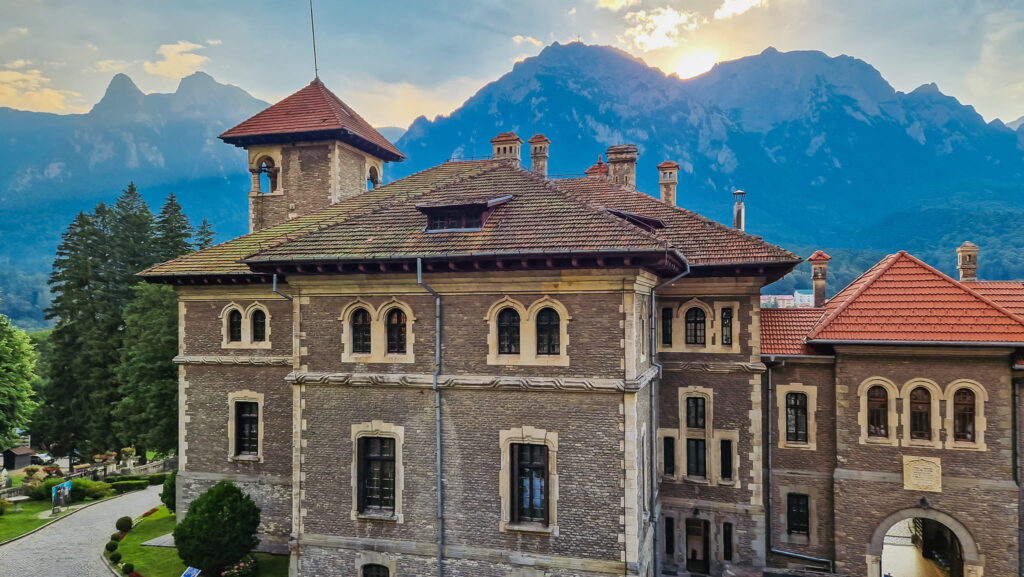 Cantacuzino Castle, aka Nevermore Academy in Wednesday - Historic stone building with red-tiled roofs and small towers, set against a backdrop of blue mountains at sunset; a cobblestone path in foreground.