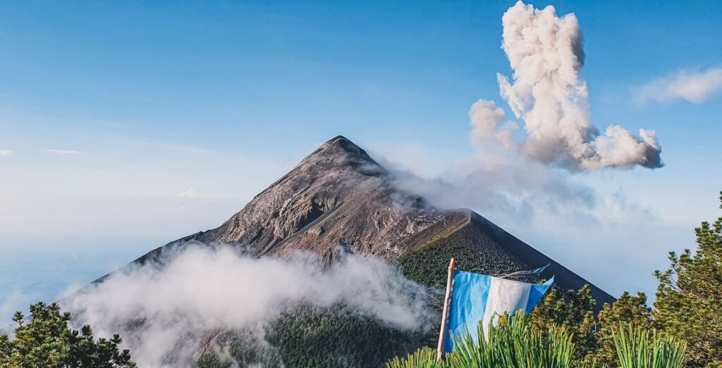 Active stratovolcano with a tall plume of white smoke against a blue sky, seen from a hillside with blue-and-white banners in the foreground.