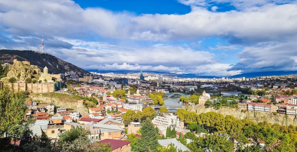 Tbilisi - panoramic cityscape with hills, a river, and a mix of historic and modern buildings under a blue, cloudy sky. A tall communications tower sits on a hill to the left.