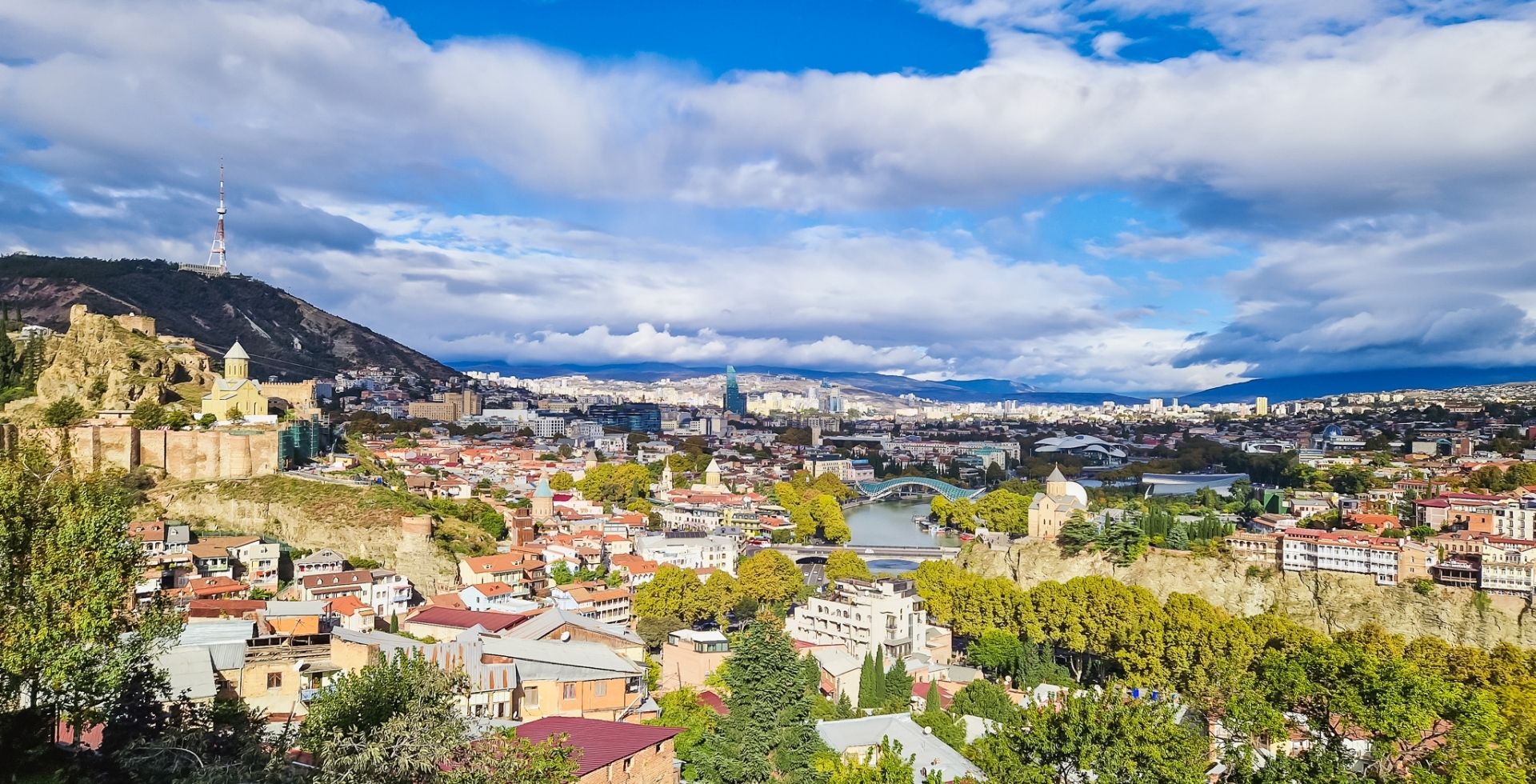 Tbilisi - panoramic cityscape with hills, a river, and a mix of historic and modern buildings under a blue, cloudy sky. A tall communications tower sits on a hill to the left.