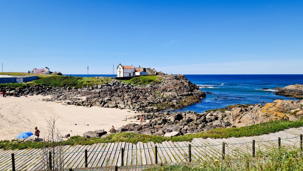 Camino Portugues Day One Coastal scene with a white chapel on rocky promontory, blue sea, and sandy beach in foreground under clear sky