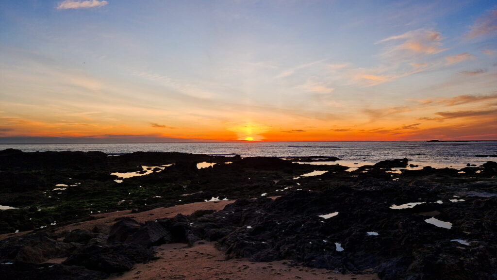 Camino Portugués Sunset Sunset over a rocky shoreline with a calm sea and orange-pink sky on the horizon.