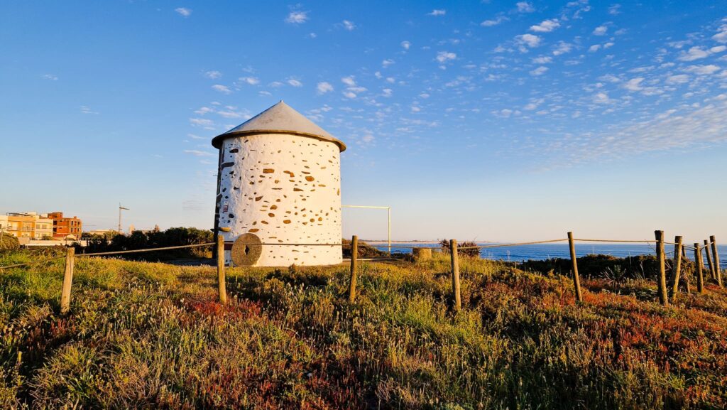 Camino Portugués - Day 2 White cylindrical stone tower with a conical roof set in a fenced grassy coastal area under a blue sky.