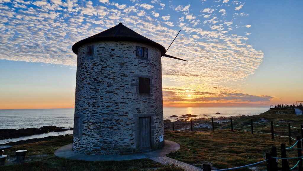 Windmill, Camino Round stone windmill by the sea at sunset, with a door, small windows, and a dramatic cloud-filled sky.