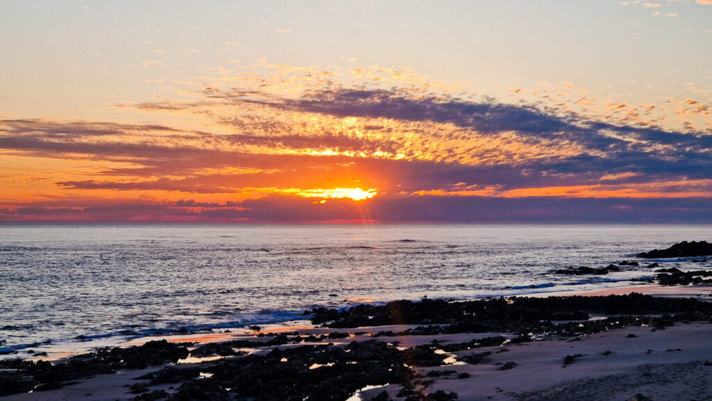 Sunset on Camino Portugués Sunset over the ocean with orange and purple clouds, rocky shoreline in foreground and calm waves.