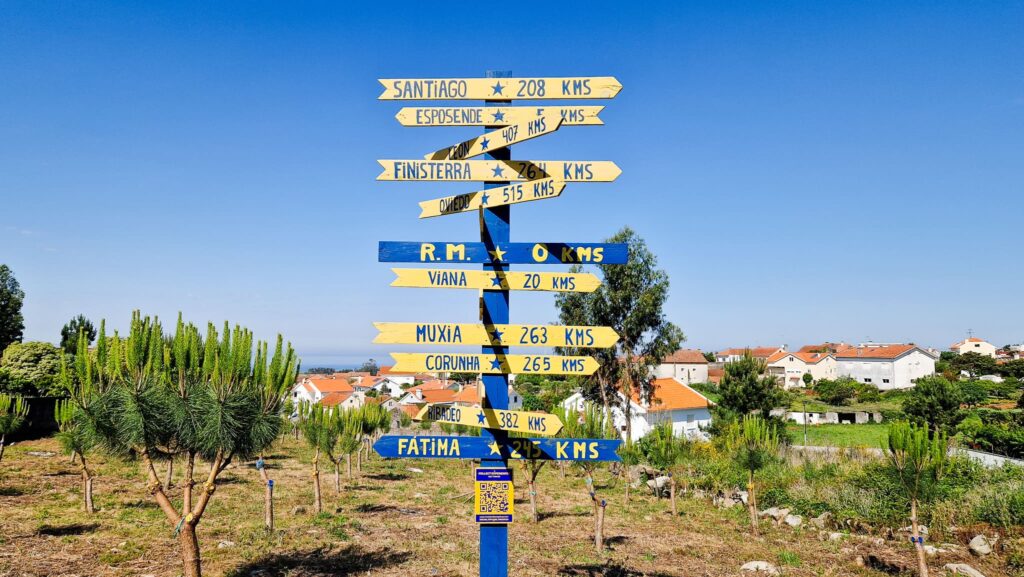 Camino Arrows Blue and yellow directional signpost with multiple arrows listing towns and distances in kms against a clear blue sky. A rural town with houses and trees in the background. How long does the Camino Portugués take?