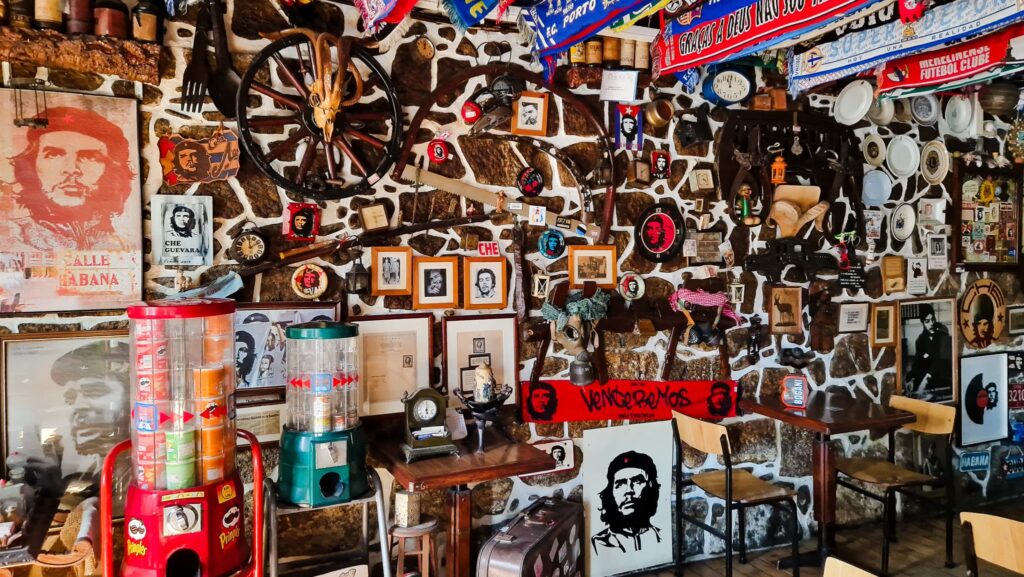 Camino Portugués Cafe Wall densely covered with Cuban memorabilia: Che Guevara posters, framed photos, clocks, and colorful scarves on a stone wall in a cozy cafe setting.
