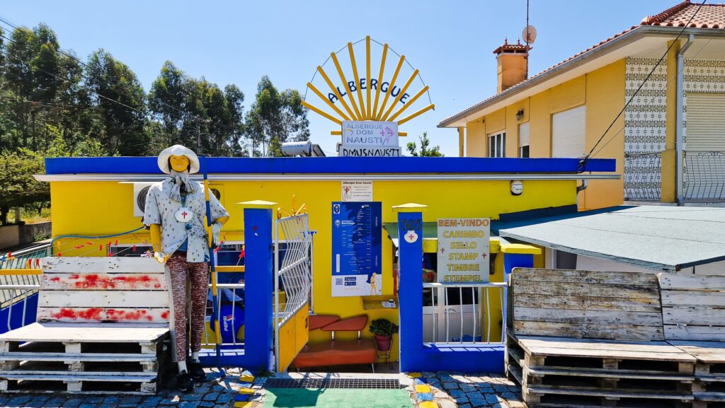 Camino Portugués Albergue Colorful yellow and blue storefront with a life‑size mannequin wearing a hat, mannequins and signs advertising stamps in Portuguese. Albergue on the Camino Portugués