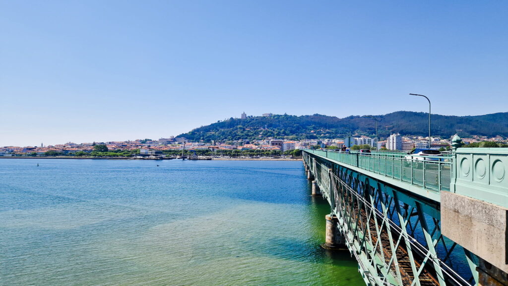 Camino Portugués - Day 3 Bridge extending over calm blue water toward a hillside town with trees and low buildings under a clear sky.