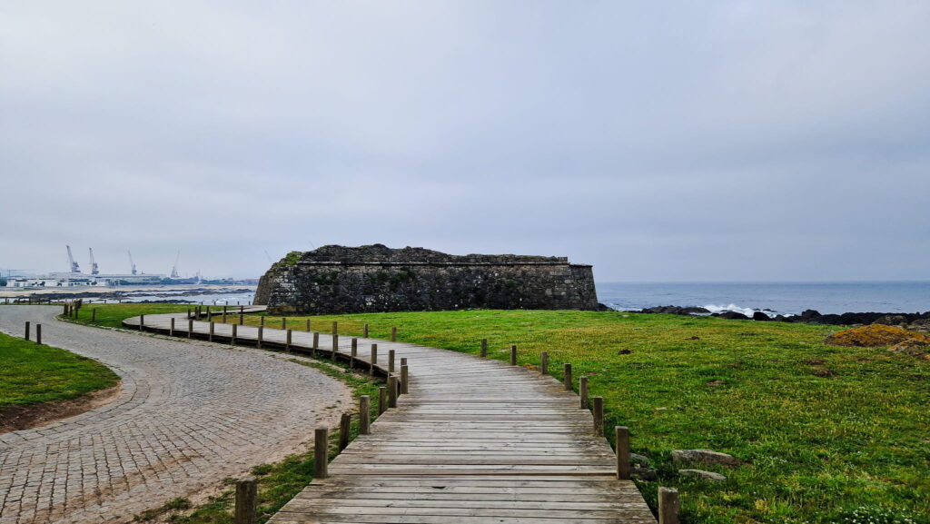 Camino Portugués - Day 4 Stone sea fort along a grassy shoreline with a curved wooden boardwalk leading to the water.