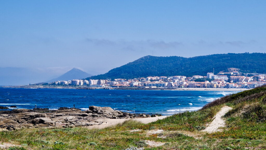 Âncora Coastal town along a rocky shoreline with a green hill in the background and blue sea in the foreground.