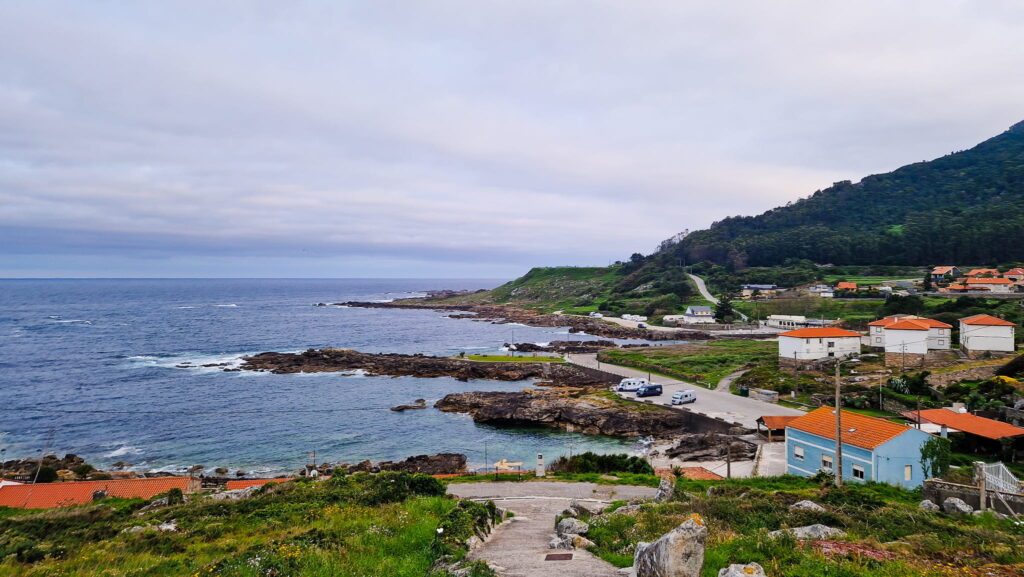 Roxky Shore, Cloudy, Camino Coastal village along rocky shore with blue sea, green hills, and orange-roofed houses perched on a hillside.