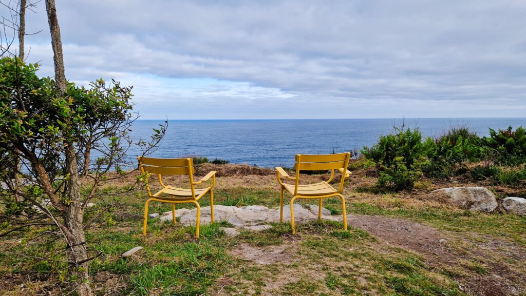 Camino Portugués - Day 5 Two yellow chairs sit on a grassy cliff overlooking a calm blue sea under a cloudy sky, with green shrubs nearby.