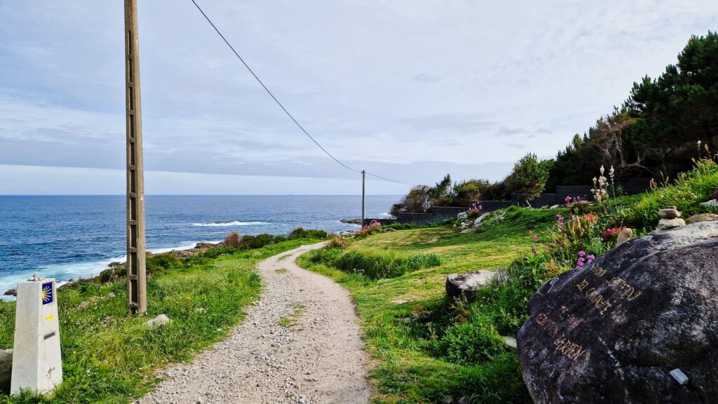 Coastal Camino, Spain Coastal gravel path runs along the sea with rocks, greenery, and a utility pole on the left under a blue sky.