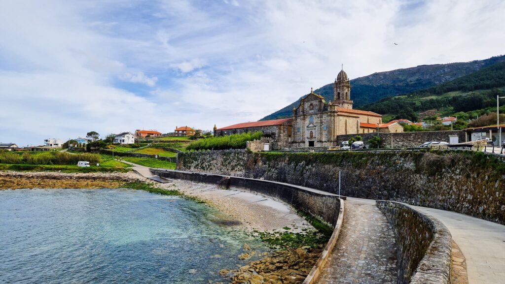 Coastal town with a historic stone church and bell tower beside a stone seawall, overlooking calm blue water and hills in the distance.