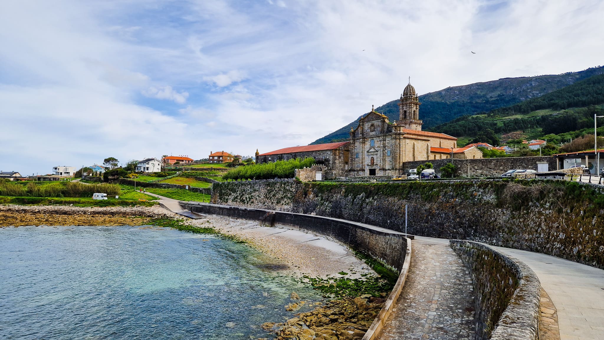 Coastal town with a historic stone church and bell tower beside a stone seawall, overlooking calm blue water and hills in the distance.