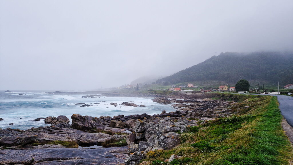 Raini day on the Camino Portugués Rocky shoreline with crashing waves in the foreground, and a misty hillside village along a coastal road in the distance.