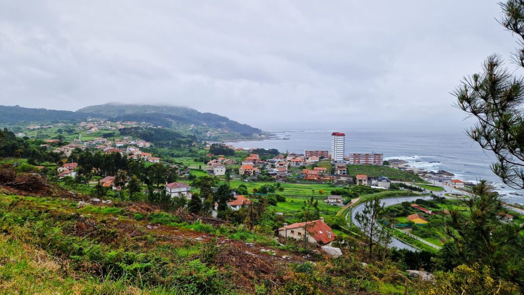 Cloudy Camino day Coastal town on green hills with a winding road, red-roof houses, and the sea under cloudy skies.