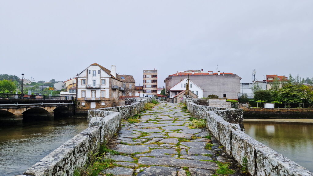 Bayona Stone stone-paved pedestrian bridge extending toward white and beige townhouses along a calm river, under a gray overcast sky.
