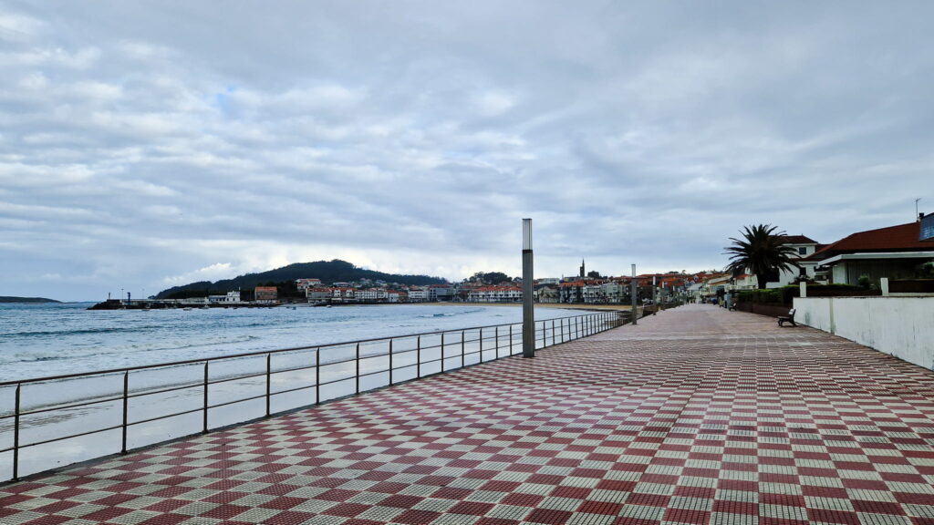 Seaside Camino Seaside promenade with red-and-white checkered tiles, railing along the water, and a coastal town in the distance under cloudy skies.