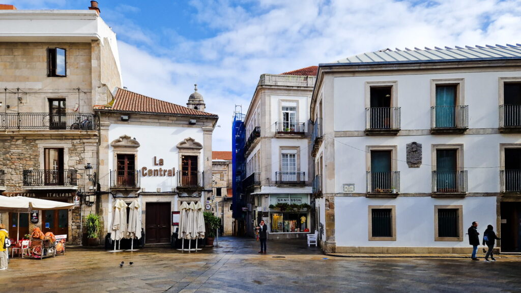 Vigo Day Off Sunlit square in Vigo, Spain, with historic stone buildings, outdoor cafe tables with white umbrellas, and pedestrians passing by the shop labeled La Central.
