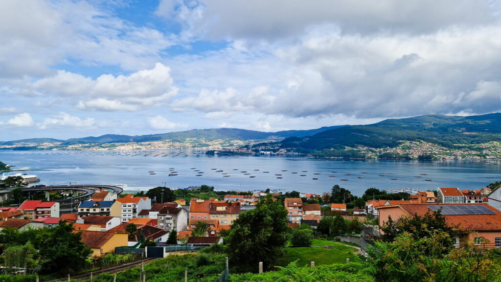 Outside Vigo Coastal town with red-tiled houses along a calm bay and green hills under a cloudy sky, boats on the water