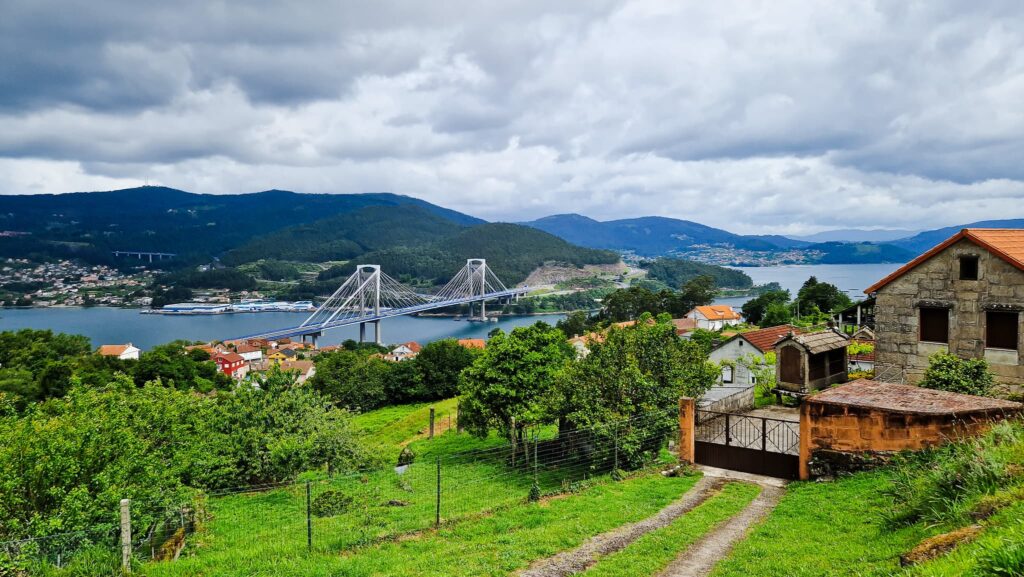 Leaving Vigo Hillside village beside a wide river with a white cable-stayed bridge and green fields under a cloudy sky