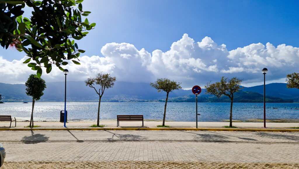 Cesantes Seafront promenade with benches, trees, and street lamps along calm water, mountains in the distance under a blue sky with clouds.