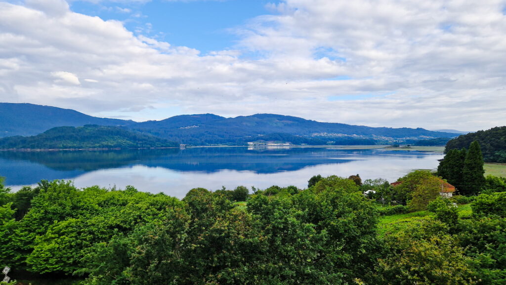 Camino rest day Wide lake reflecting blue sky and surrounding green hills, with forested foreground and distant mountains under scattered clouds