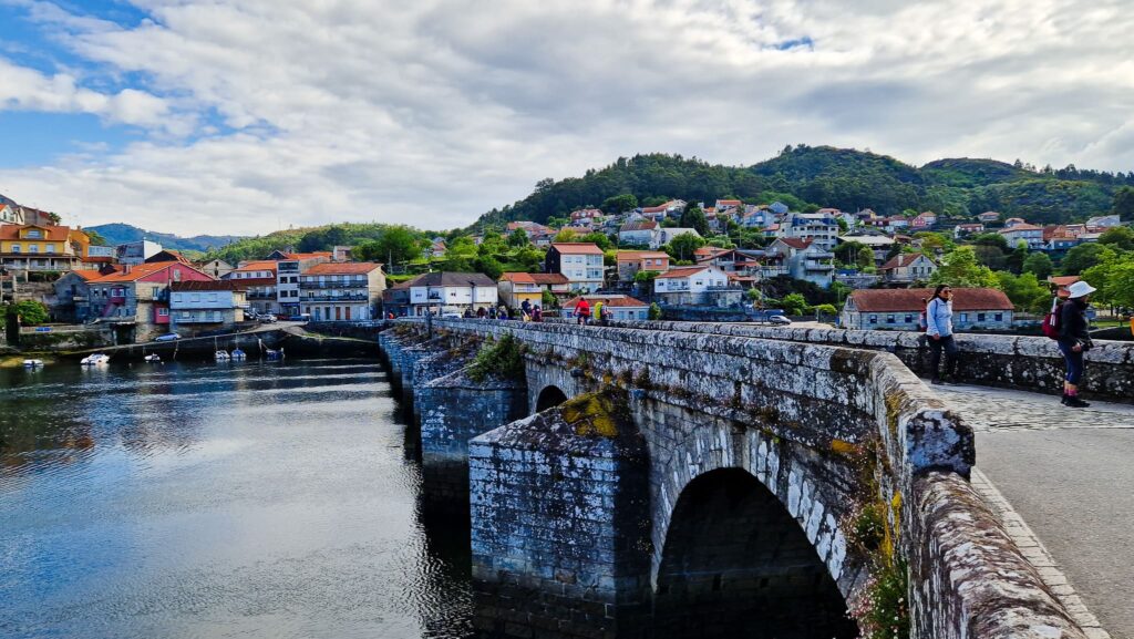Arcade Stone arched bridge over a harbor with colorful hillside houses and people walking along the quay under a cloudy sky.