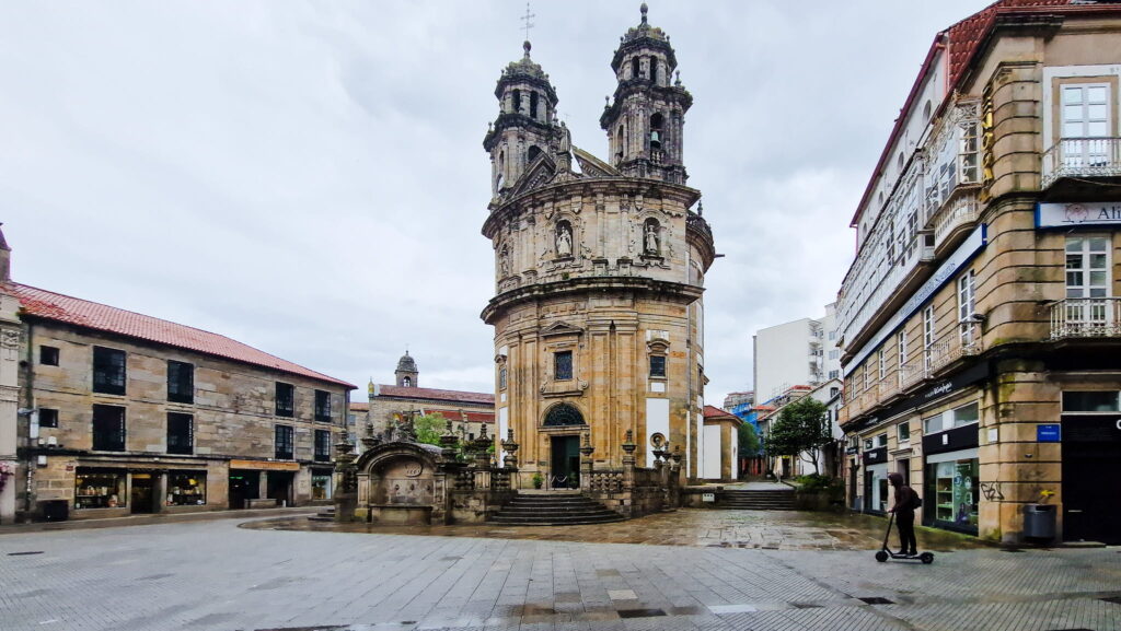 Pontevedra Historic stone church with two ornate towers in a square in Pontevedra, surrounded by shops and pedestrians, including a person on a scooter in the foreground.