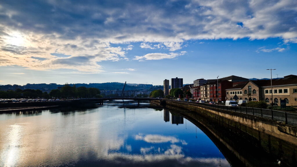 Camino Portugués - Stage 10 Riverside cityscape with buildings on the right, a bridge in the distance, and a calm river reflecting the cloudy sky and blue water.