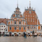 Riga, Latvia ornate red-brick town hall-style building with white decorative trim and tall sculpted gables in a cobblestone square; people walking and horse carts nearby, overcast sky