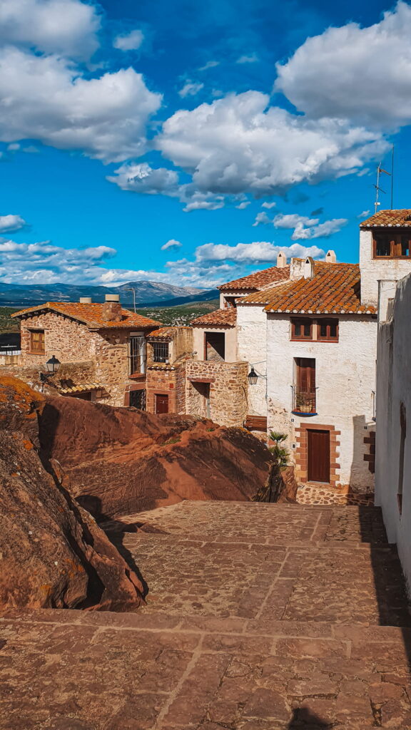Vilafamés Sunny hillside village with white stone houses and red-tiled roofs along a cobblestone street under a bright blue sky with clouds.