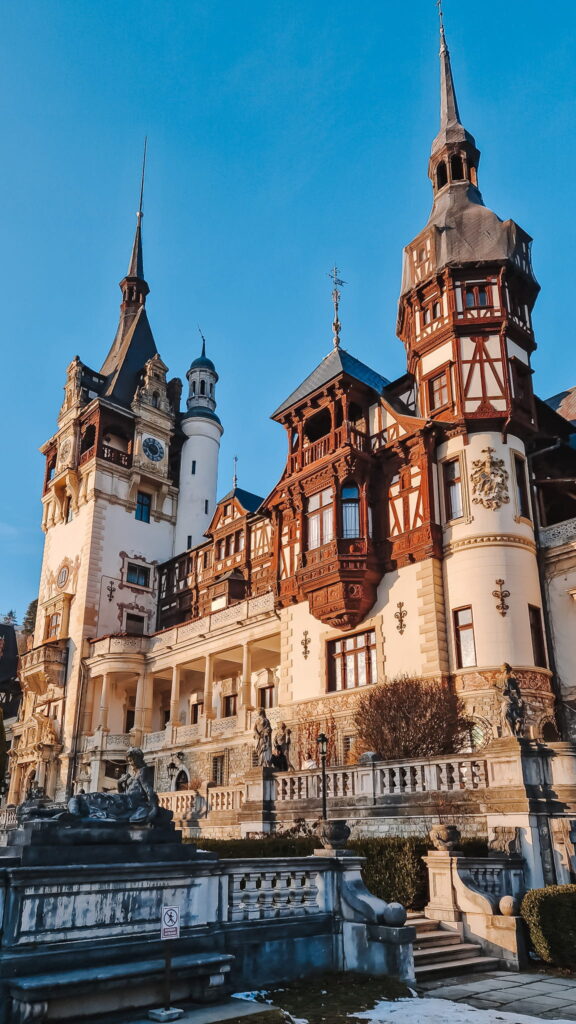 20231210_152052 Ornate European castle with tall spires, red-brick and beige stone, statues in the foreground against a blue sky.