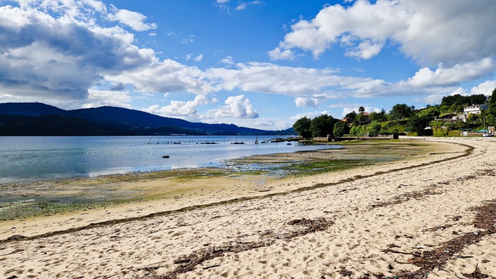 Camino Portugués - Cesantes Wide sandy beach along calm blue water with hills in the distance and houses on a hillside to the right under a partly cloudy sky, coastal scene.