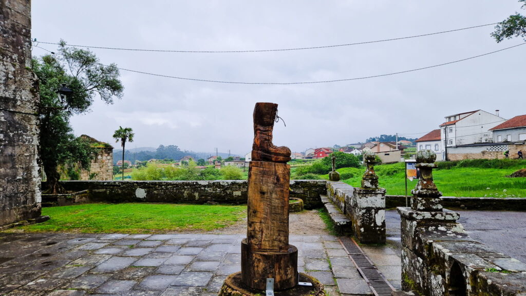 Camino Portugués - Boot Carved wooden boot sculpture on a pedestal in a stone courtyard, with green grass and distant houses under cloudy skies.