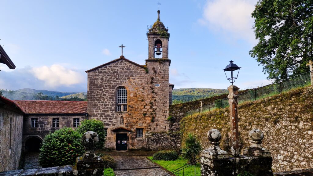 Herbon, Camino Portugués Stone church with a bell tower in a walled courtyard, blue sky and distant hills behind it.