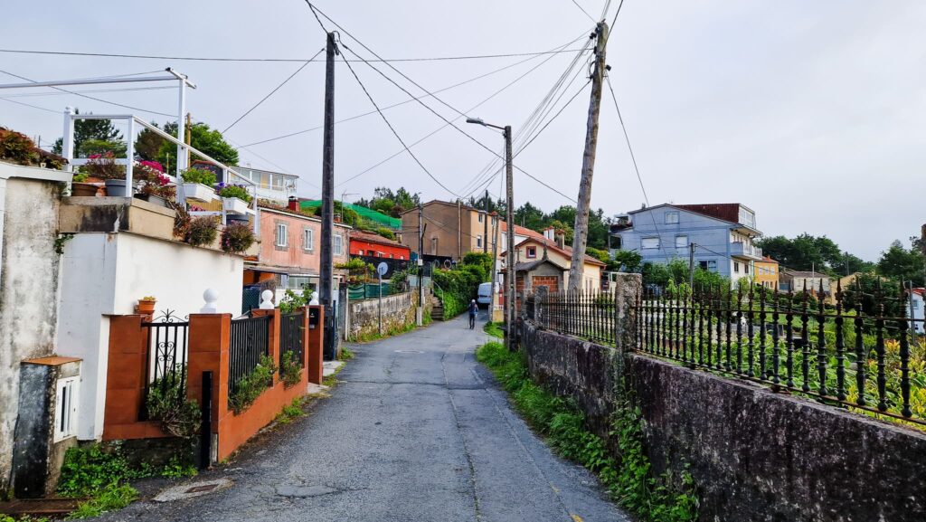 Camino Portugués - Day 13 Residential hill street with colorful houses, potted plants, and overhead power lines; a cyclist in the distance under a pale sky.