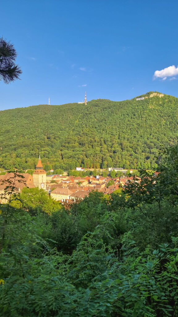 20240715_191927 Forested hill above a town with red-roofed houses in Romania; a church steeple rises near the bottom left under a clear blue sky.