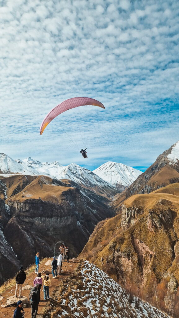 Georgia mountains Paraglider with pink wing soaring above snow-capped mountains as spectators watch from a rocky trail, Georgia