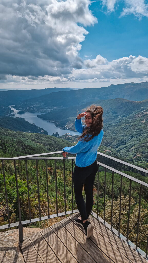 Rox in Portugal Woman in a blue sweater stands on a metal railing at a scenic overlook, with a valley, river, and mountains in the distance. Portugal travel