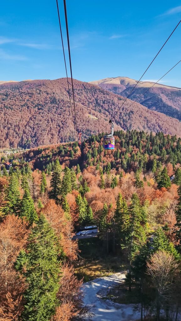 20251101_135756 Cable car suspended above a forested valley with autumn-colored trees and distant mountains under a blue sky; passengers ride in the car. Romania travel