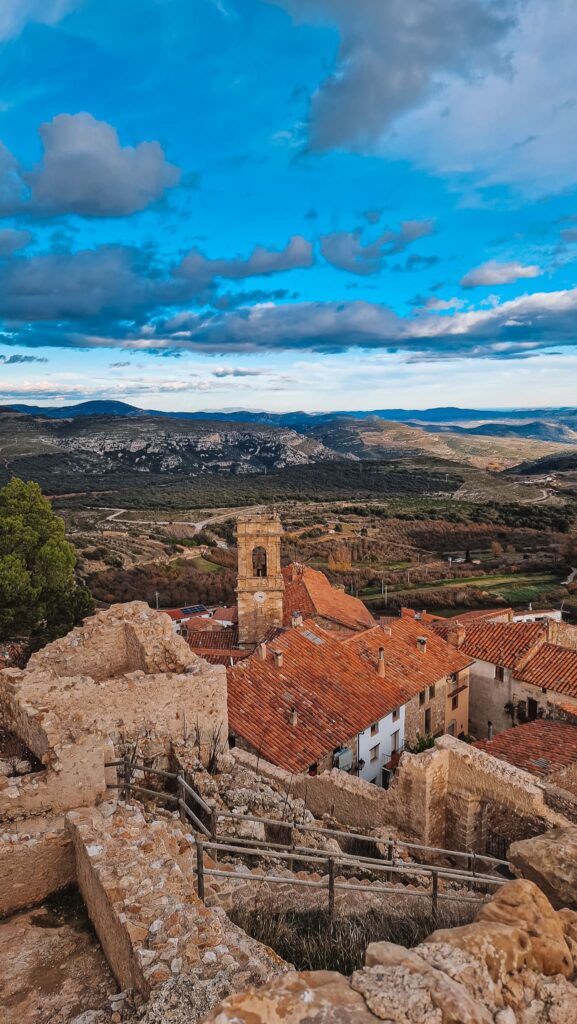 Culla Ruined stone walls and red-tiled roofs of a hillside village with a bell tower under a blue, cloudy sky.