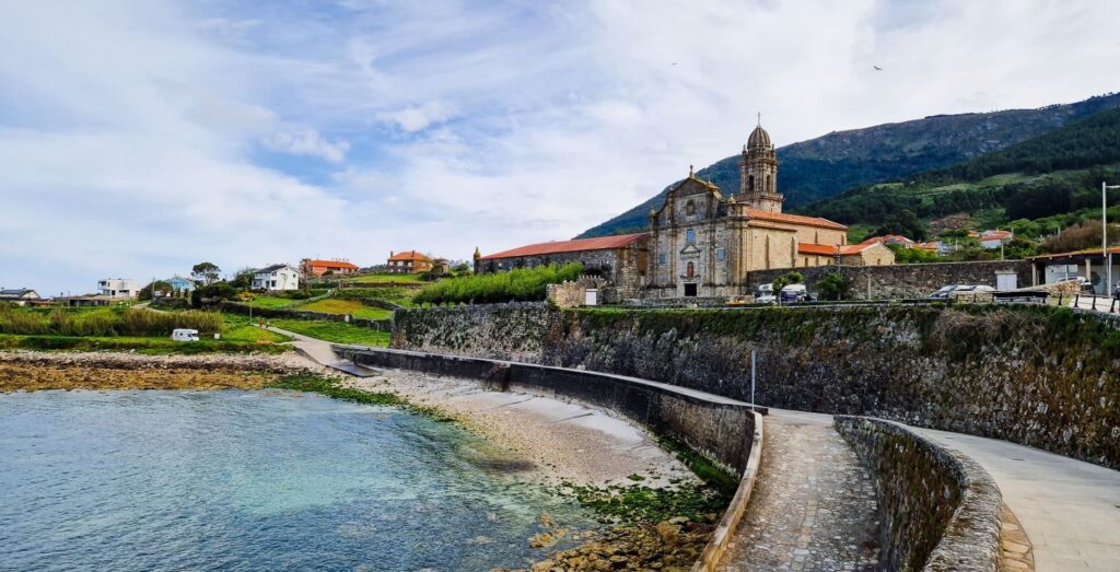 Coastal village with a stone church and bell tower along a curved seawall, sea in foreground and hillside houses beyond.
