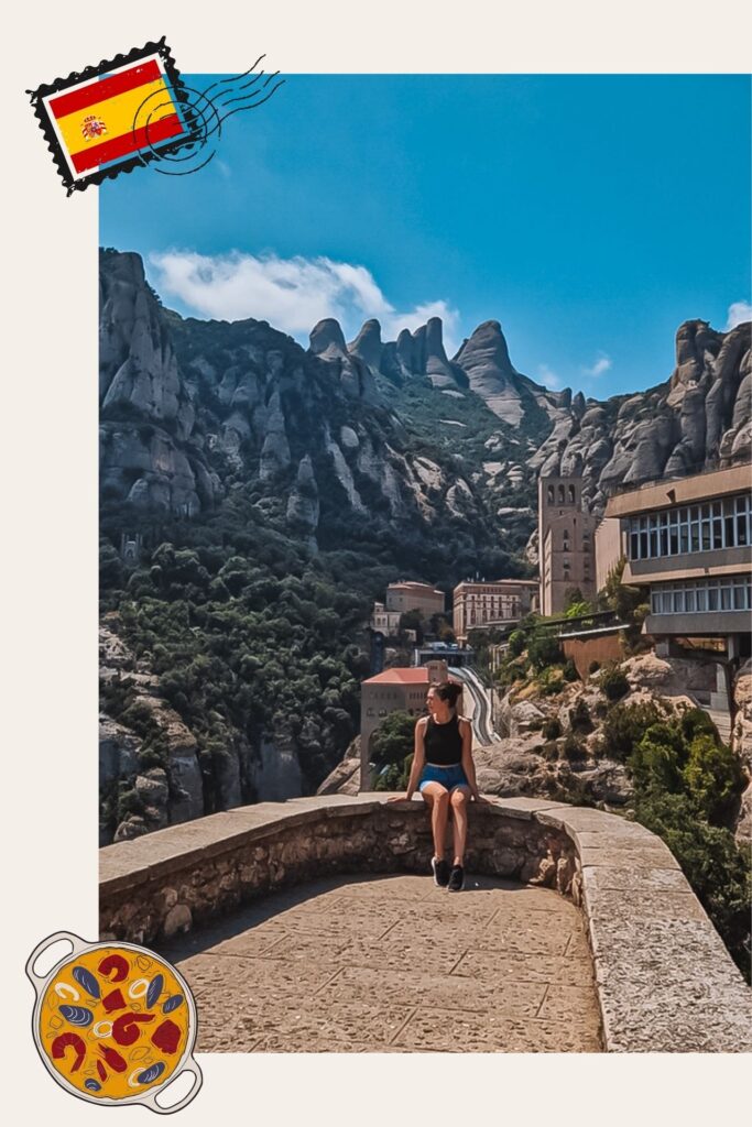 RoxInSpain Woman sits on a stone railing along a sunny mountain path with jagged rock formations and buildings in the background.