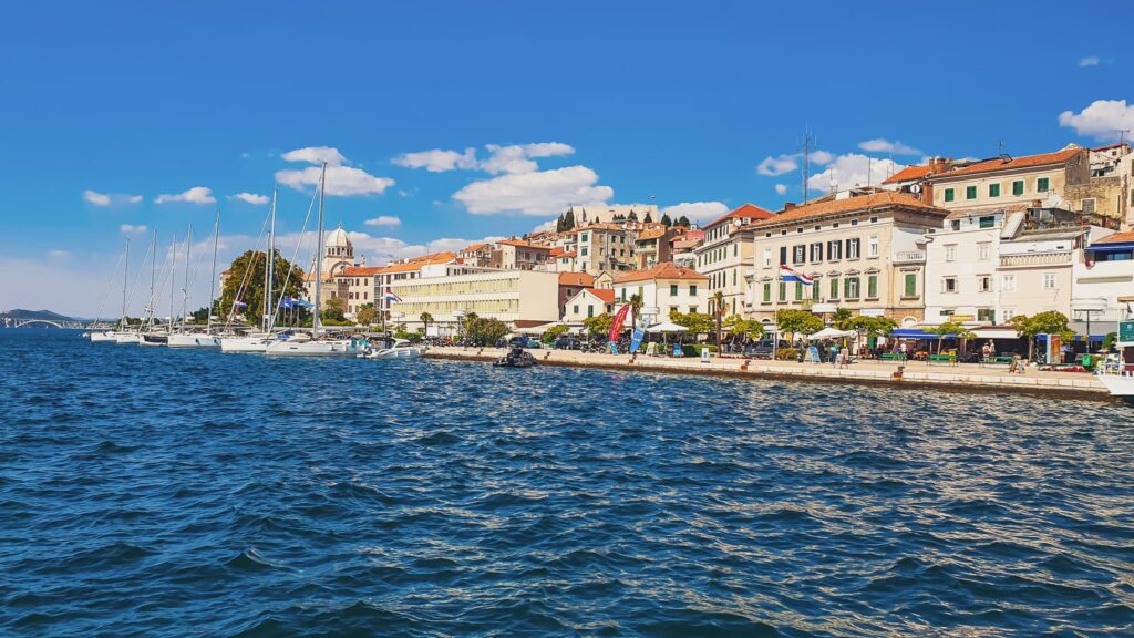 Šibenik town view Harbor lined with sailboats and a row of red-roofed buildings along the water under a clear blue sky.