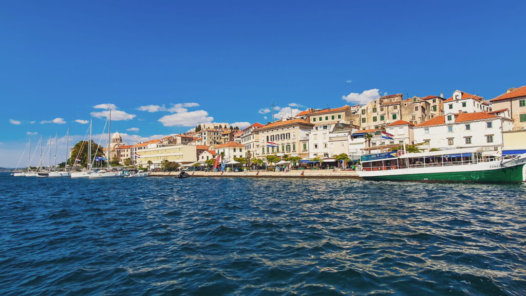 Šibenik seaside town Harbor town with white buildings and red-tiled roofs along the shore, sailboats moored in the foreground under a blue sky.